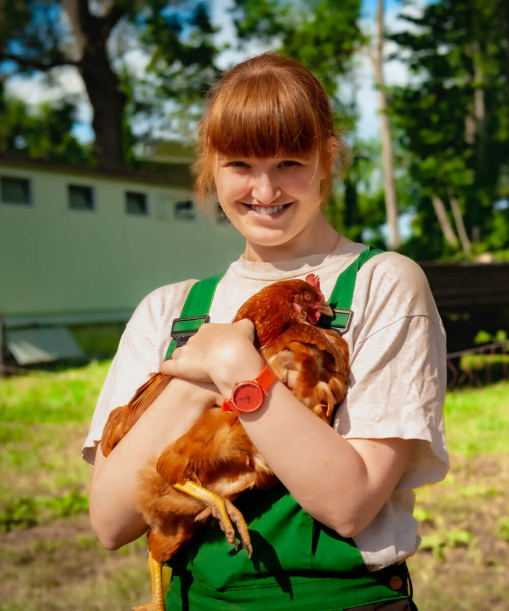 Eine Mitarbeiterin in Arbeitskleidung steht auf der Wiese und hält ein Huhn im Arm.